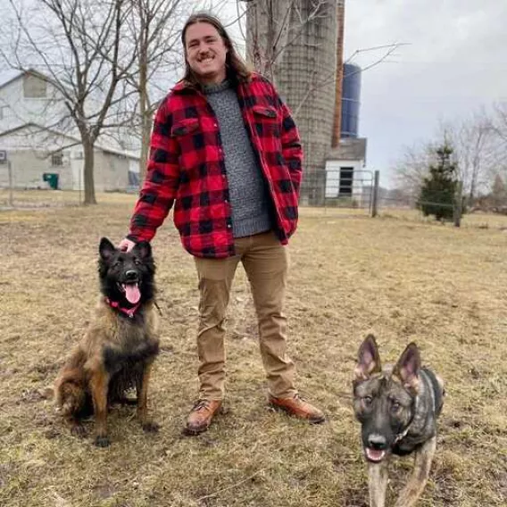 A man with brown hair on a farm, in autumn clothes, standing with two dogs.